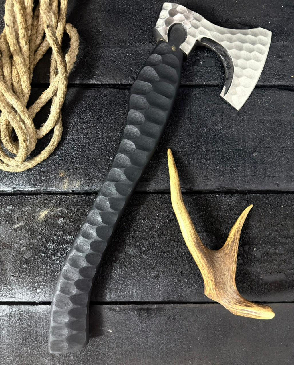 Axes with textured handles on a dark wooden surface, accompanied by a coiled rope and deer antler.