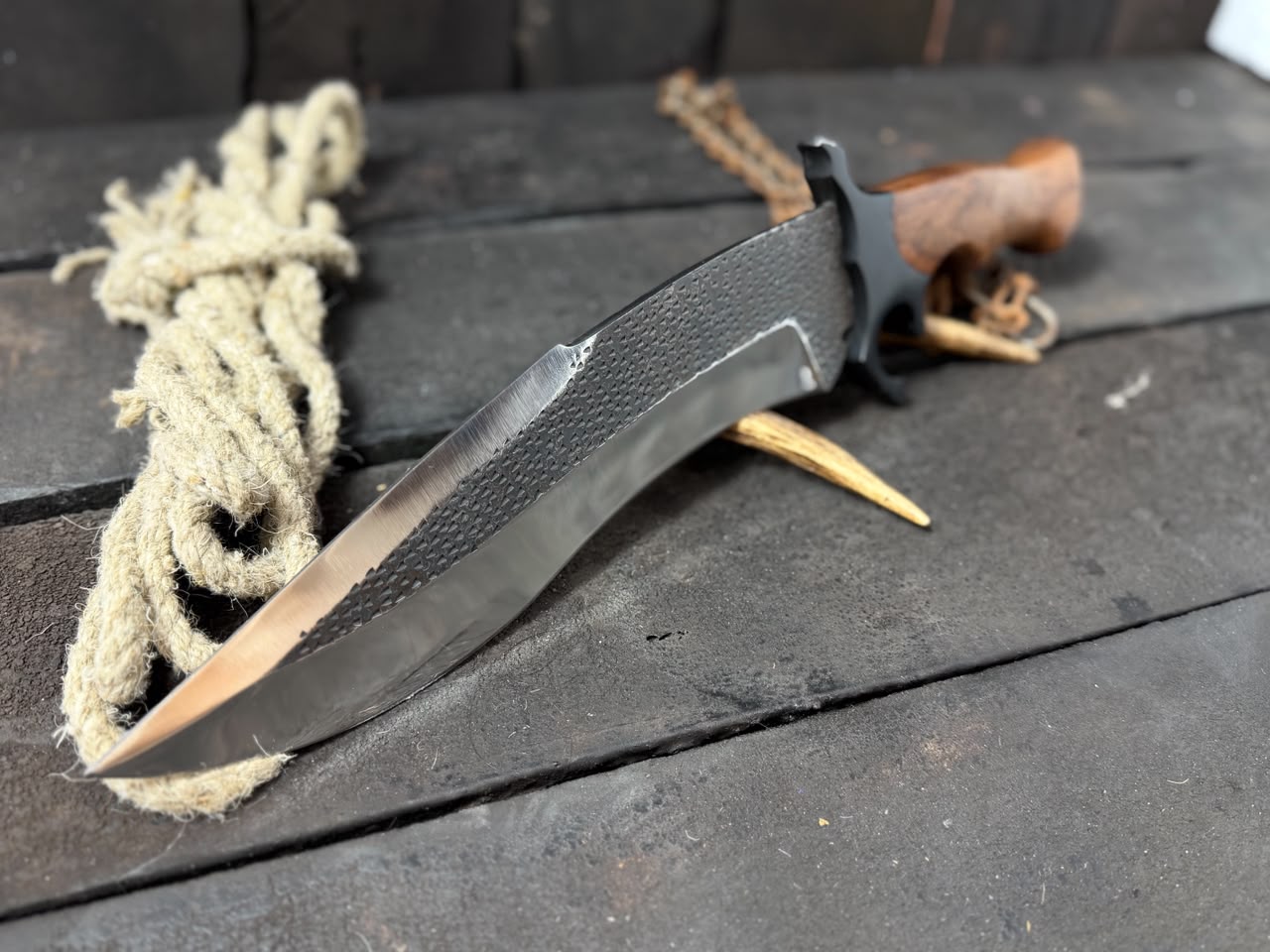 Knife with a textured black handle and blade on a wooden surface with rope and wood pieces.