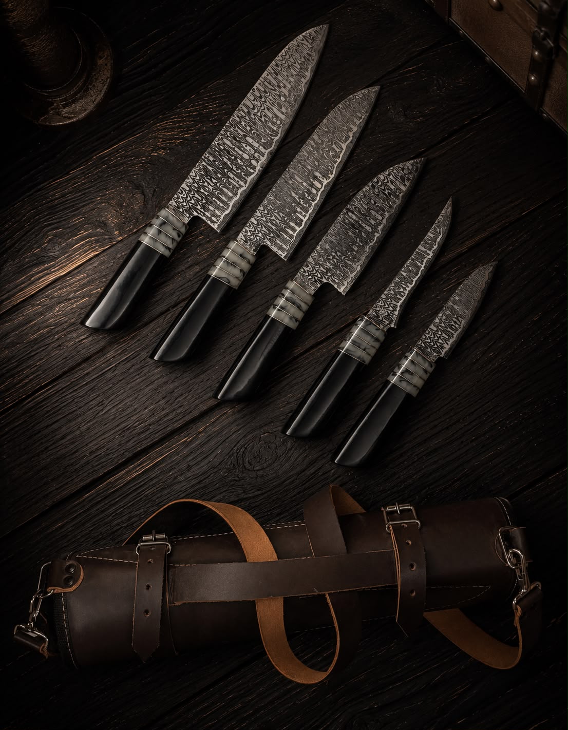Set of four Damascus steel knives with black handles on a dark wooden surface, accompanied by a leather knife sheath.