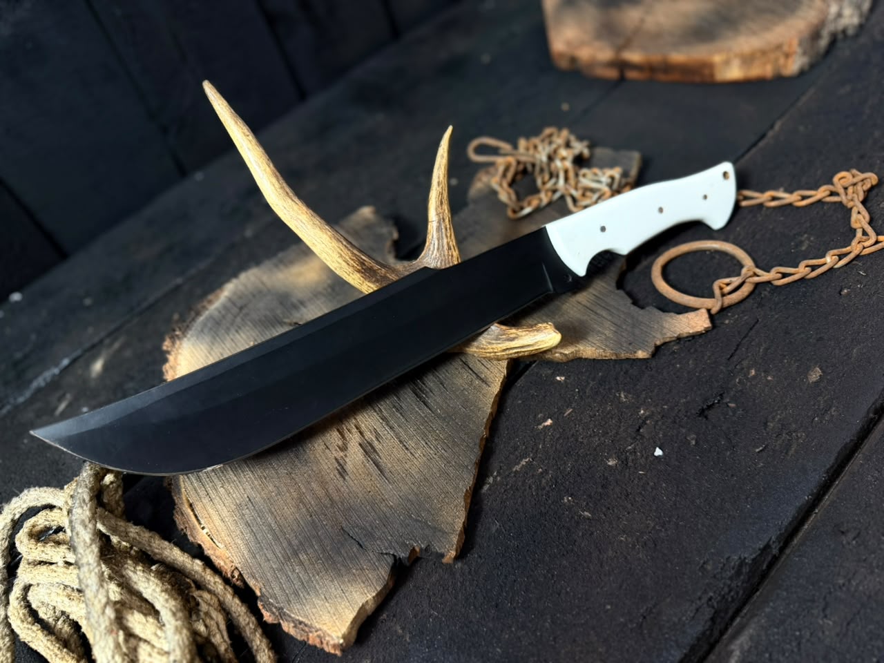 Knife with a black blade and white handle on a wooden surface with antlers and chains.