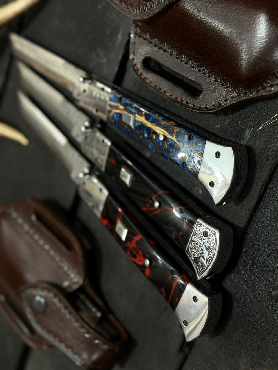 Three folding knives with decorative handles in a leather sheath on a dark surface.