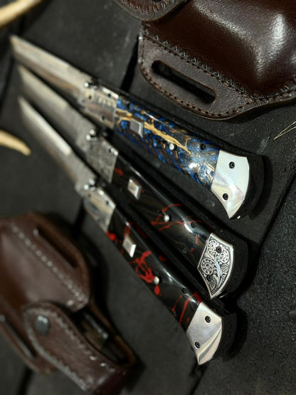 Three folding knives with decorative handles in a leather sheath on a dark surface.