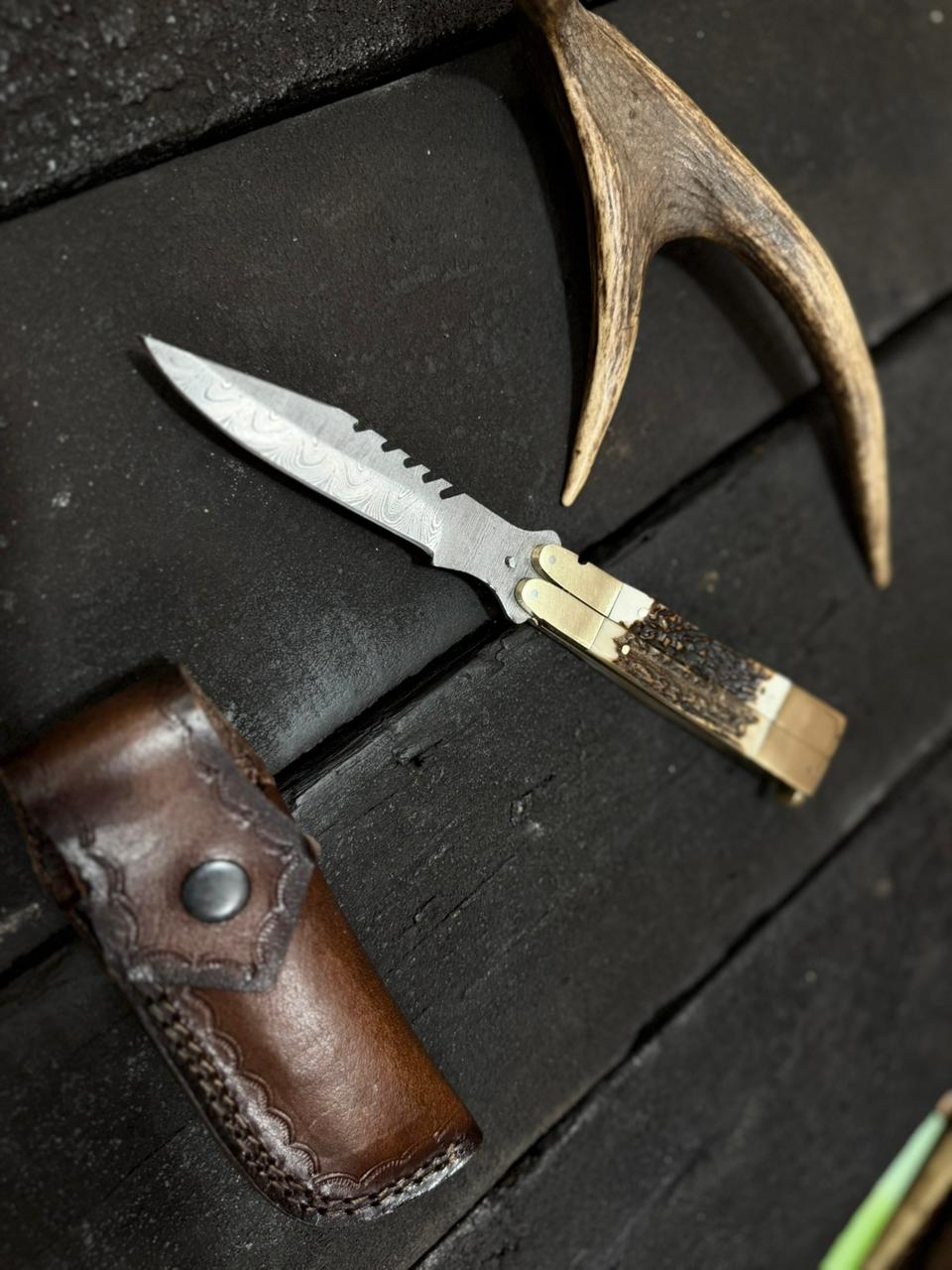 Damascus Butterfly Knife with leather sheath and antler on a dark wooden surface