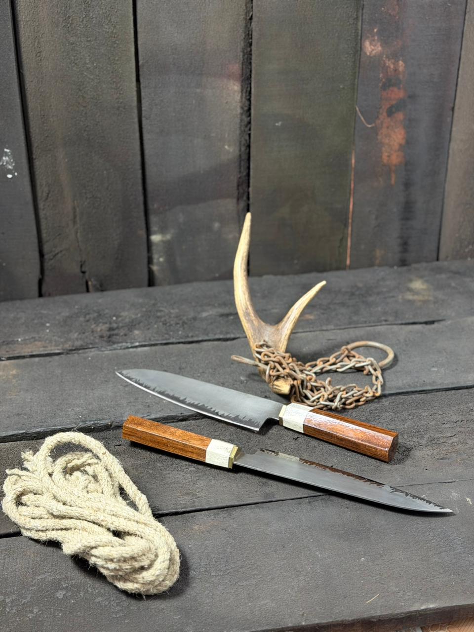 Two knives with wooden handles and metal blades on a dark wooden surface, accompanied by a rope and deer antlers.