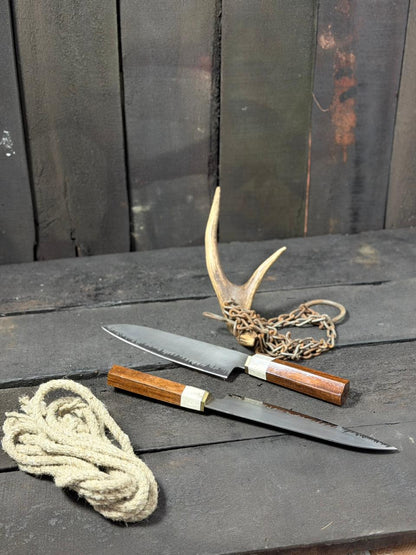 Two knives with wooden handles and metal blades on a dark wooden surface, accompanied by a rope and deer antlers.